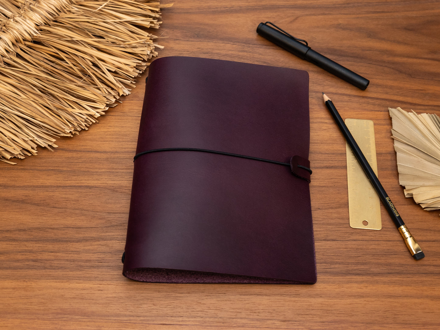 A dark purple leather journal with a black elastic closure strap, displayed on a wooden desk alongside stationery items including a gold pen, pencils, a ruler, and a woven straw mat.