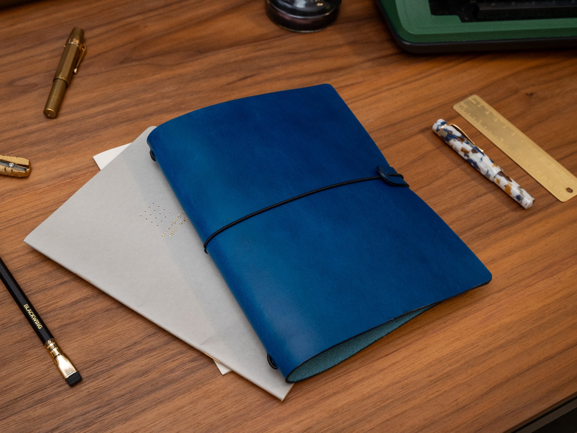 A dark blue leather journal with a black elastic closure strap, displayed on a wooden desk alongside stationery items including a gold pen, pencils, a ruler, and a woven straw mat.