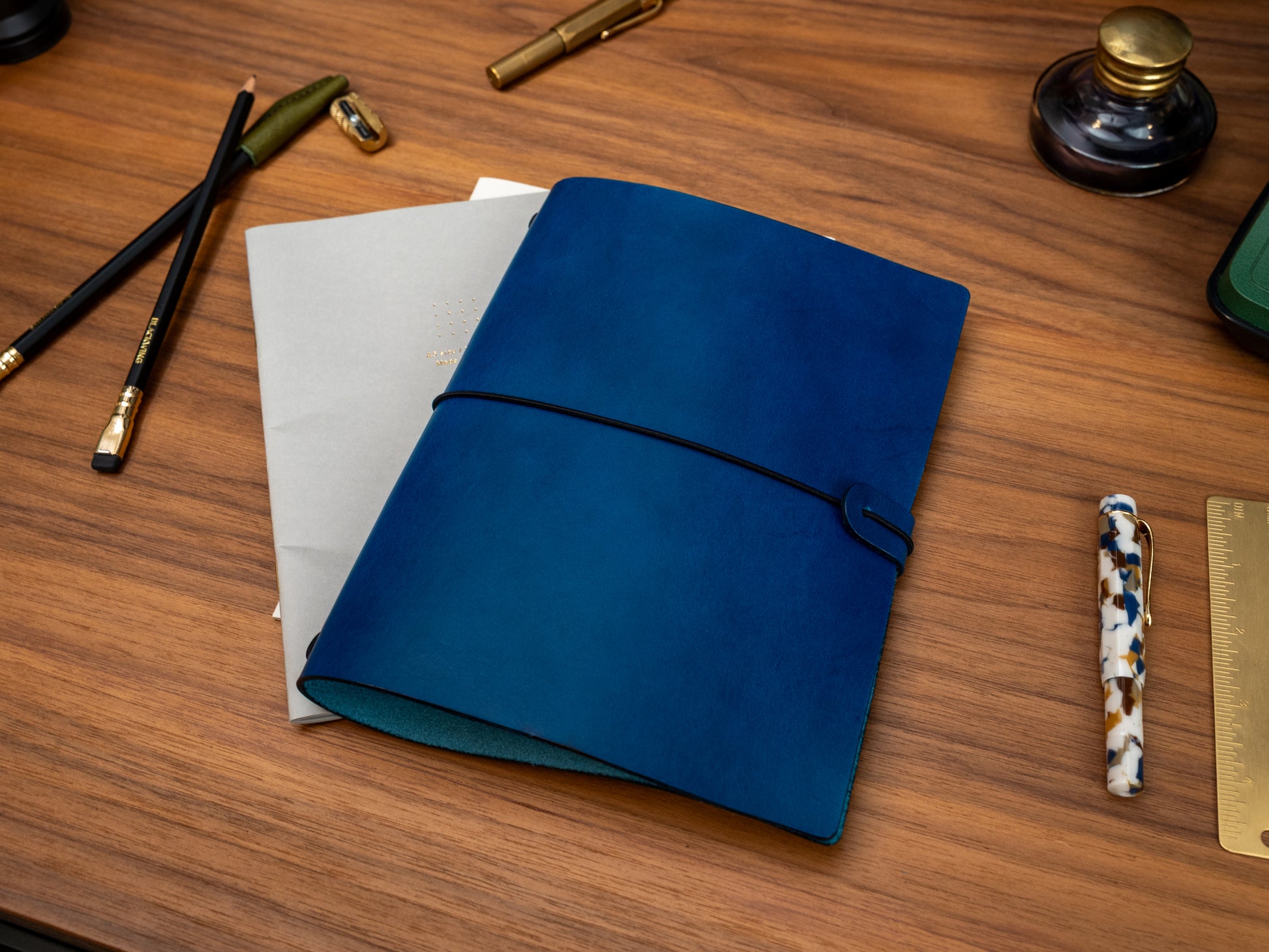 A dark blue leather journal with a black elastic closure strap, displayed on a wooden desk alongside stationery items including a gold pen, pencils, a ruler, and a woven straw mat.