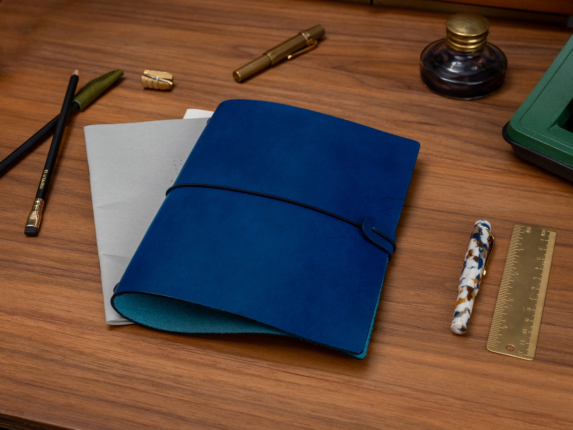 A dark blue leather journal with a black elastic closure strap, displayed on a wooden desk alongside stationery items including a gold pen, pencils, a ruler, and a woven straw mat.