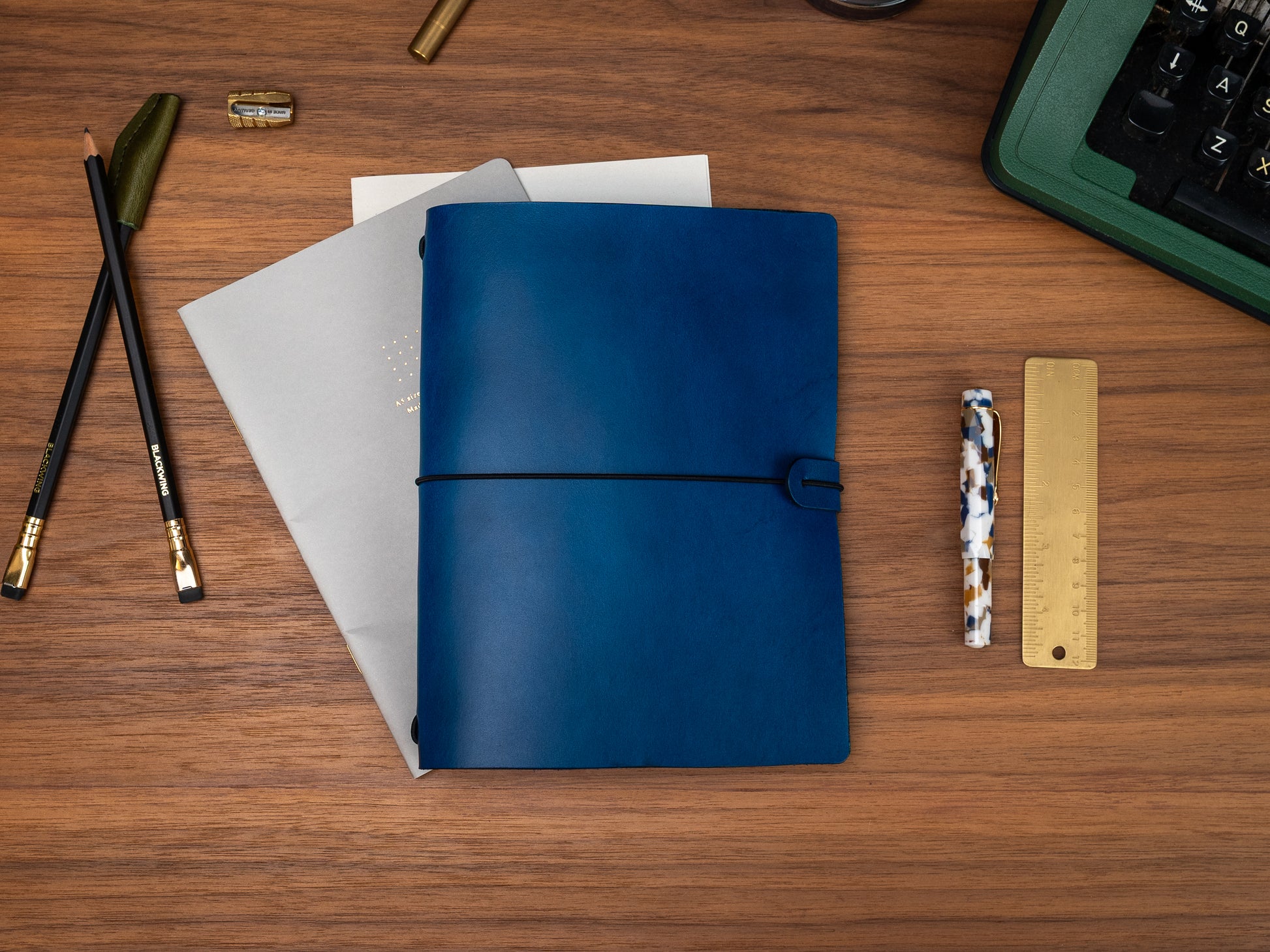 A dark blue leather journal with a black elastic closure strap, displayed on a wooden desk alongside stationery items including a gold pen, pencils, a ruler, and a woven straw mat.